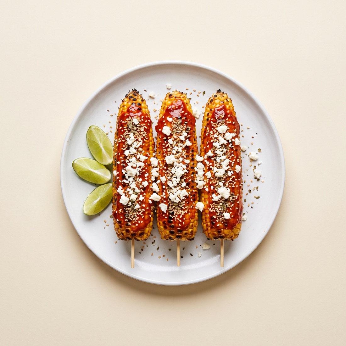 Tres elotes asados glaseados con gochujang, queso fresco y orégano sobre un plato blanco, vista cenital